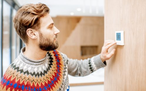 Man in sweater feeling cold adjusting room temperature with electronic thermostat at home